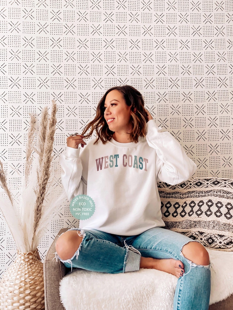 Puede incluir: Una sudadera blanca con el texto "WEST COAST" en letras de colores. La sudadera est&aacute; hecha con tintas a base de agua, es ecol&oacute;gica, no t&oacute;xica y sin PVC.