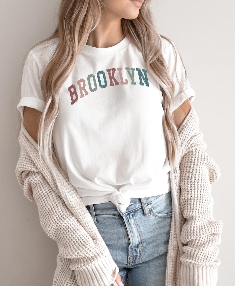 Op de afbeelding: Wit T-shirt met het woord "BROOKLYN" in een regenboogvormig ontwerp. Het shirt is in de taille geknoopt en wordt gedragen met een blauwe jeans en een cr&egrave;mekleurige cardigan.
