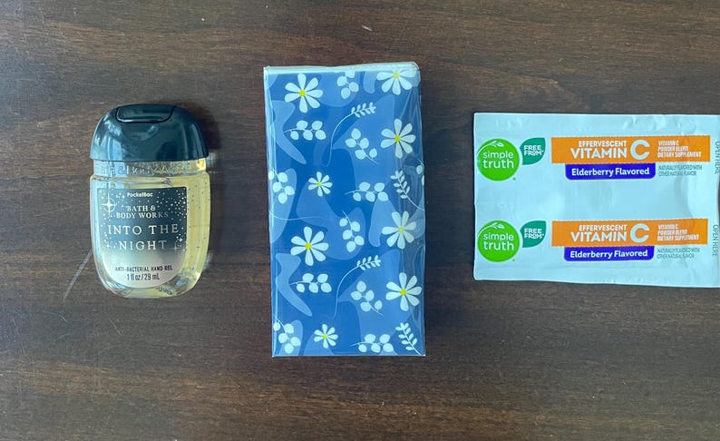 May include: A Bath & Body Works Into The Night anti-bacterial hand gel in a 1 fl oz bottle, a pack of tissues with a blue and white floral pattern, and two Simple Truth effervescent Vitamin C elderberry flavored dietary supplement packets.