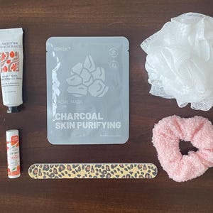 May include: A white tube of hand cream, a gray facial mask packet labeled Charcoal Skin Purifying, a white mesh bath sponge, a pink textured scrunchie, a small tube of mango lip balm, and a long nail file with a brown and black leopard print pattern.