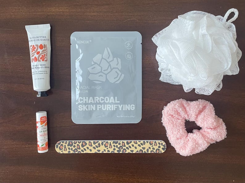 May include: A white tube of hand cream, a gray facial mask packet labeled Charcoal Skin Purifying, a white mesh bath sponge, a pink textured scrunchie, a small tube of mango lip balm, and a long nail file with a brown and black leopard print pattern.