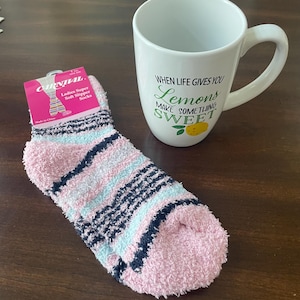 May include: A white ceramic mug with the text "WHEN LIFE GIVES YOU Lemons MAKE SOMETHING SWEET" and a yellow lemon graphic, paired with a fuzzy ankle slipper sock featuring pink, light blue, and dark navy blue horizontal stripes and a pink tag.