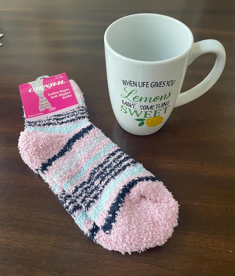 May include: A white ceramic mug with the text "WHEN LIFE GIVES YOU Lemons MAKE SOMETHING SWEET" and a yellow lemon graphic, paired with a fuzzy ankle slipper sock featuring pink, light blue, and dark navy blue horizontal stripes and a pink tag.