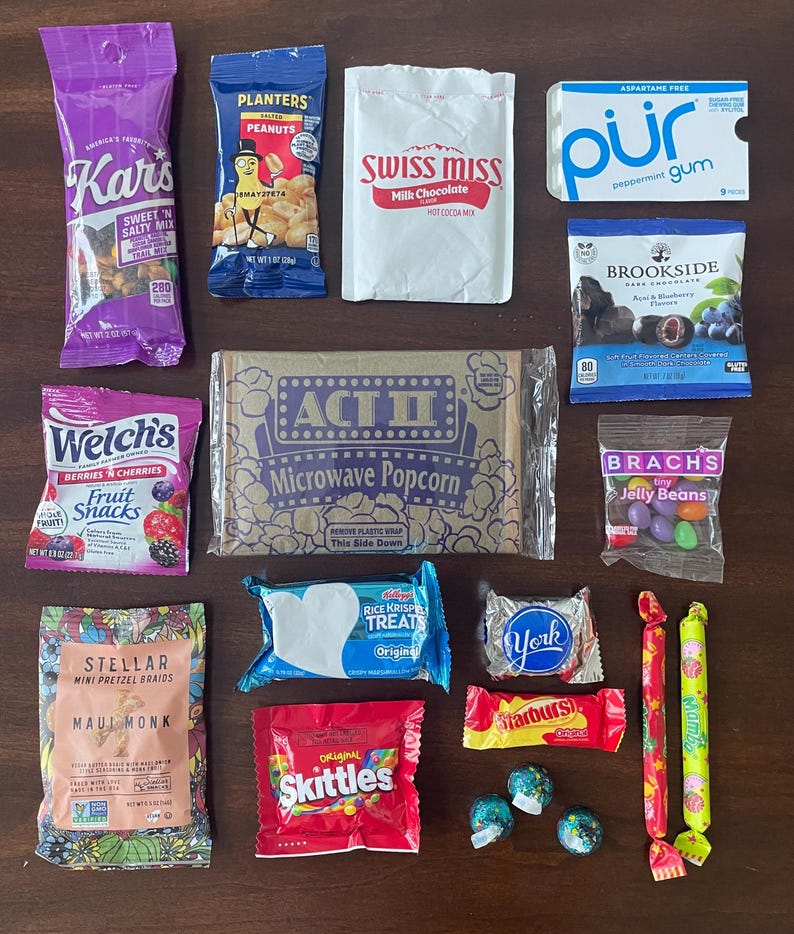 May include: A collection of various snack food packages including Kar's Sweet 'n Salty Mix, Planters Salted Peanuts, Swiss Miss Hot Cocoa, P&uuml;r Peppermint Gum, Brookside Dark Chocolate, Welch's Fruit Snacks, Act II Microwave Popcorn, Brach's Jelly Beans, Stellar Mini Pretzel Braids, Rice Krispies Treats, York Peppermint Pattie, Starburst, Skittles, and two Mamba fruit chews with three small round candies.