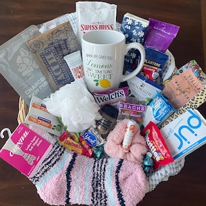 May include: A gift basket containing a white ceramic mug with the text When Life Gives You Lemons Make Something Sweet, pink and blue striped slipper socks, a white bath sponge, a pink scrunchie, facial masks, and various snacks including popcorn, chocolate, candy, and gum.