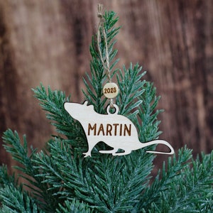 May include: A wooden Christmas bauble shaped like a rat with the name "MARTIN" and the year "2023" on it. The bauble is hanging on a green artificial Christmas tree.