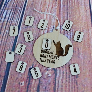 Può includere: Un ornamento natalizio in legno a forma di sfera con una silhouette nera di un gatto. L'ornamento ha il testo "BROKEN ORNAMENTS THIS YEAR" e un set di etichette numerate da 0 a 10.