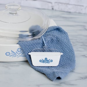 Könnte beinhalten: Weiße Auflaufformen mit blauen Blumenmustern. Eine kleine rechteckige Schale liegt auf einem blauen Handtuch. Eine größere Auflaufform mit Glasdeckel befindet sich im Hintergrund. Die Schalen stehen auf einer Marmoroberfläche.