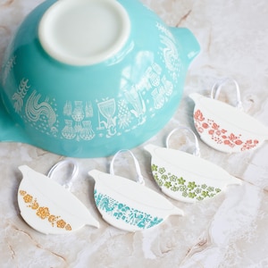 Puede incluir: Cuatro adornos de cerámica blanca con forma de cuencos con diferentes patrones florales vintage. Los adornos cuelgan de una cinta blanca y están sobre una superficie gris claro. Un cuenco turquesa más grande con un patrón floral blanco está en el fondo.