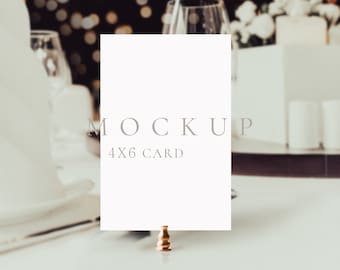 Può includere: Un mockup di una carta bianca 4x6 su un supporto dorato, con uno sfondo sfocato di una mise en place con tovaglioli bianchi e candele.
