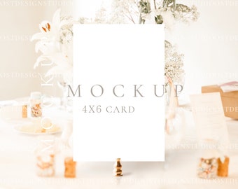 May include: A white 4x6 card mockup is displayed on a gold stand. The word "MOCKUP" is printed above the card, and "4X6 CARD" is printed below. The background includes flowers and other decorative elements, creating a soft, neutral aesthetic.