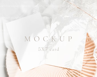May include: Two blank white 5x7 cards on a textured beige surface with a white feather branch in the background. The text "MOCKUP 5X7 card" is printed on the cards.
