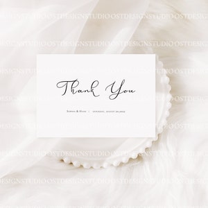May include: A white thank you card with black calligraphy that reads "Thank You" and the text "Sophia & Hans | Saturday, August 30, 2023". The card is on a white fluffy surface.