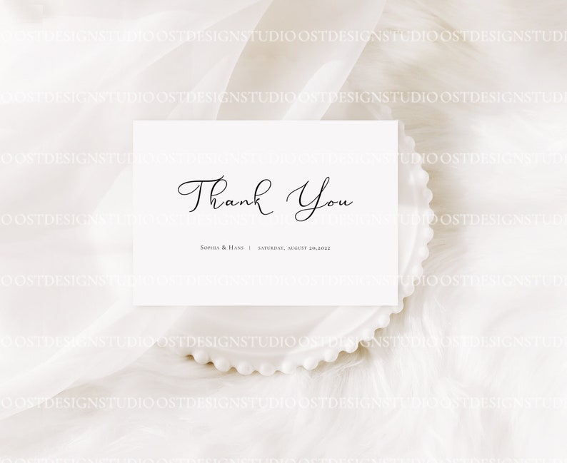 May include: A white thank you card with black calligraphy that reads "Thank You" and the text "Sophia & Hans | Saturday, August 30, 2023". The card is on a white fluffy surface.