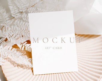 Puede incluir: Una maqueta de tarjeta blanca sobre una superficie texturizada marrón claro con ramas de helecho blancas. La tarjeta mide 5x7 pulgadas y tiene la palabra "MOCKUP" y "5x7" CARD impresa.
