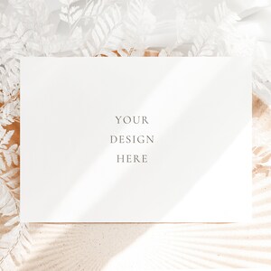 May include: A white card with the text "YOUR DESIGN HERE" on a background of white sand and white dried ferns.