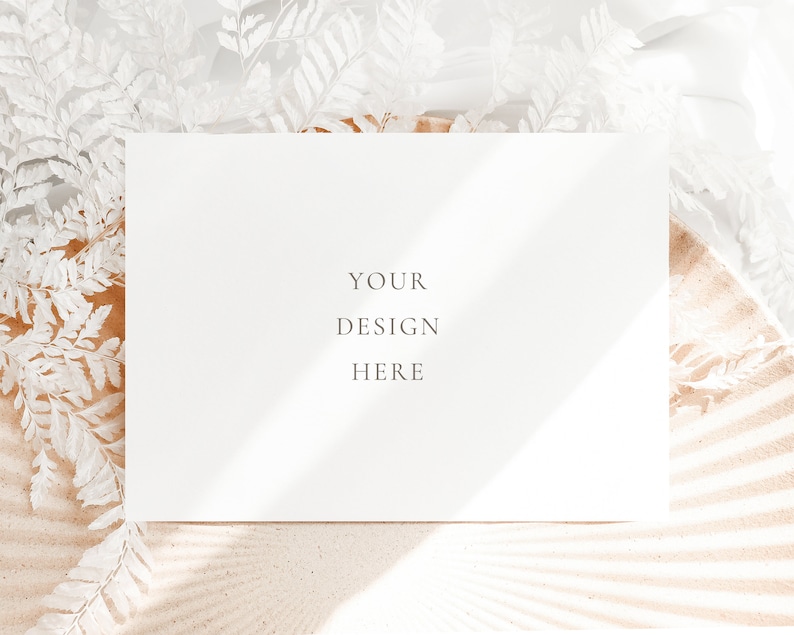 May include: A white card with the text "YOUR DESIGN HERE" on a background of white sand and white dried ferns.