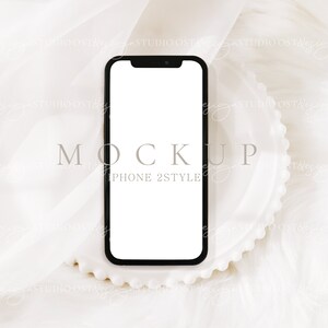 May include: A black iPhone 2 style mockup phone with a white screen, sitting on a white plate with a scalloped edge. The phone is on a white fabric background.