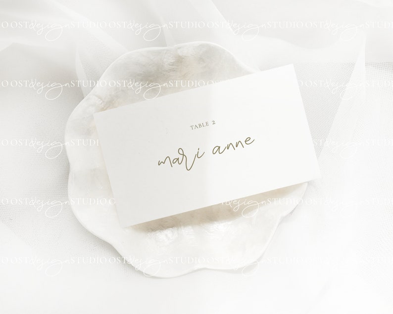 May include: White card with gold text on a white background. The card says "Table 2" and "mari anne".