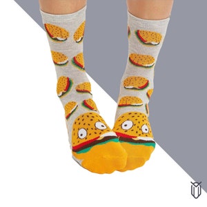 May include: A pair of grey socks with a fun burger pattern. The socks feature a yellow burger with green lettuce, red tomato, and brown bun. The burger has two eyes and a smiling mouth.