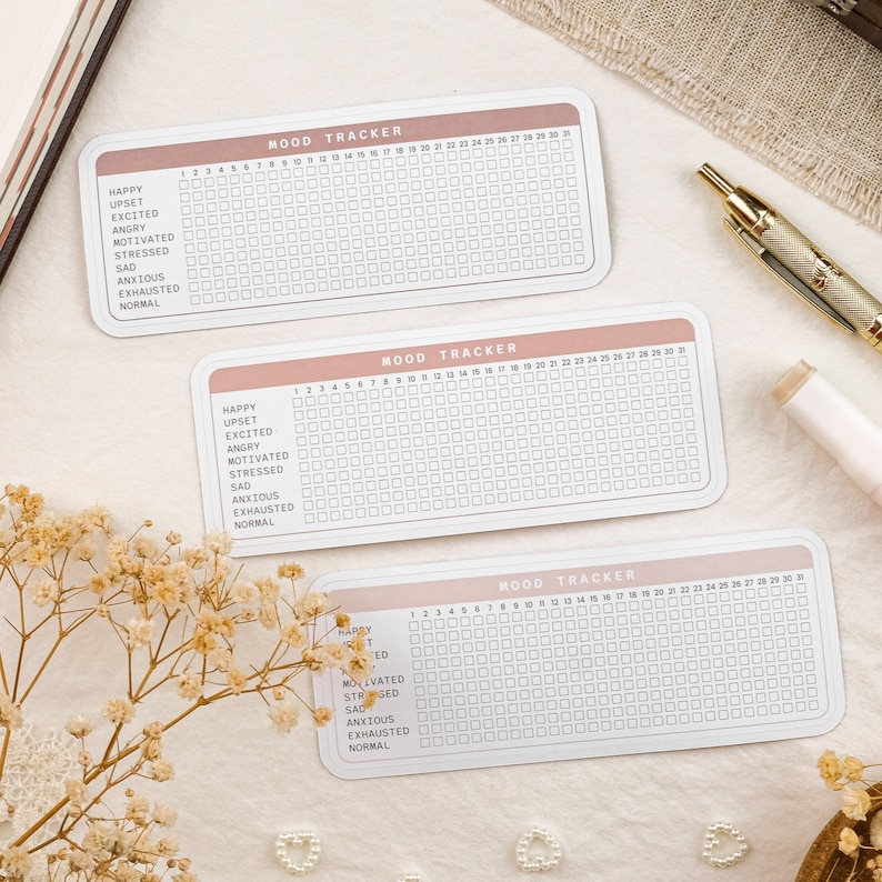 May include: Three pink and white mood tracker cards with a grid of squares and a list of moods: Happy, Upset, Excited, Angry, Motivated, Stressed, Sad, Anxious, Exhausted, Normal. The cards are on a white surface.