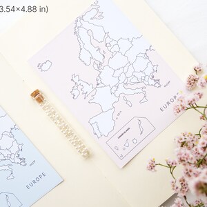 May include: Two maps of Europe, each measuring 3.54 x 4.88 inches, are displayed. One map is light blue, the other is pink. A glass vial filled with small, clear beads sits between the maps. Pink flowers are in the foreground.