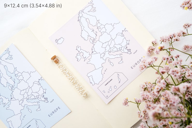 May include: Two maps of Europe, each measuring 3.54 x 4.88 inches, are displayed. One map is light blue, the other is pink. A glass vial filled with small, clear beads sits between the maps. Pink flowers are in the foreground.