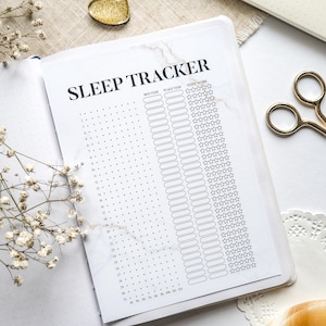 May include: Open journal with a "Sleep Tracker" page, featuring columns for bed time, wake time, and sleep score. The journal has a marble-like design. Gold scissors and a gold heart-shaped object are also visible.