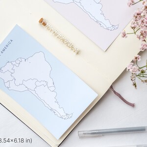 May include: A notebook with a light blue card that reads "SOUTH AMERICA" with a map outline. A small glass vial filled with small white beads and a pen are also on the notebook. The notebook is open, and a pink flower arrangement is in the background.