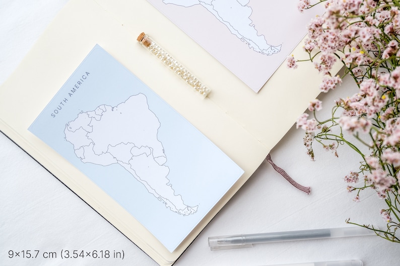 May include: A notebook with a light blue card that reads "SOUTH AMERICA" with a map outline. A small glass vial filled with small white beads and a pen are also on the notebook. The notebook is open, and a pink flower arrangement is in the background.