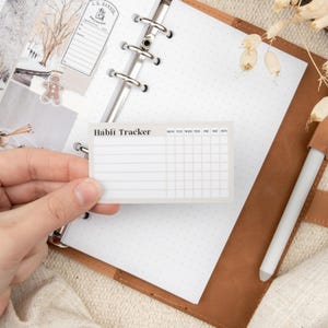 Puede incluir: Una pegatina blanca de seguimiento de hábitos con el texto "Habit Tracker" y los días de la semana etiquetados en la parte superior. La pegatina se está sujetando con una mano y se coloca en una agenda de cuero marrón.