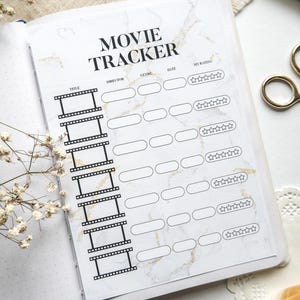 Könnte beinhalten: Offenes Filmtagebuch mit Marmordesign. Das Tagebuch hat einen Titelbereich, Regisseur, Genre, Datum und Bewertungsbereich. Die Worte "Movie Tracker" sind in Schwarz gedruckt. Das Tagebuch ist auf einer weißen Oberfläche geöffnet.
