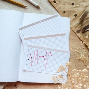 May include: A notebook with step tracker cards. The cards feature a grid and the text "STEP TRACKER". One card shows a red line graph. Pencils, star-shaped confetti, and dried flowers are also visible in the image.