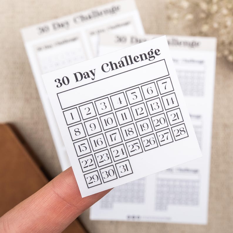May include: A white 30 Day Challenge calendar with black numbers in a grid format. The calendar is on a beige surface.