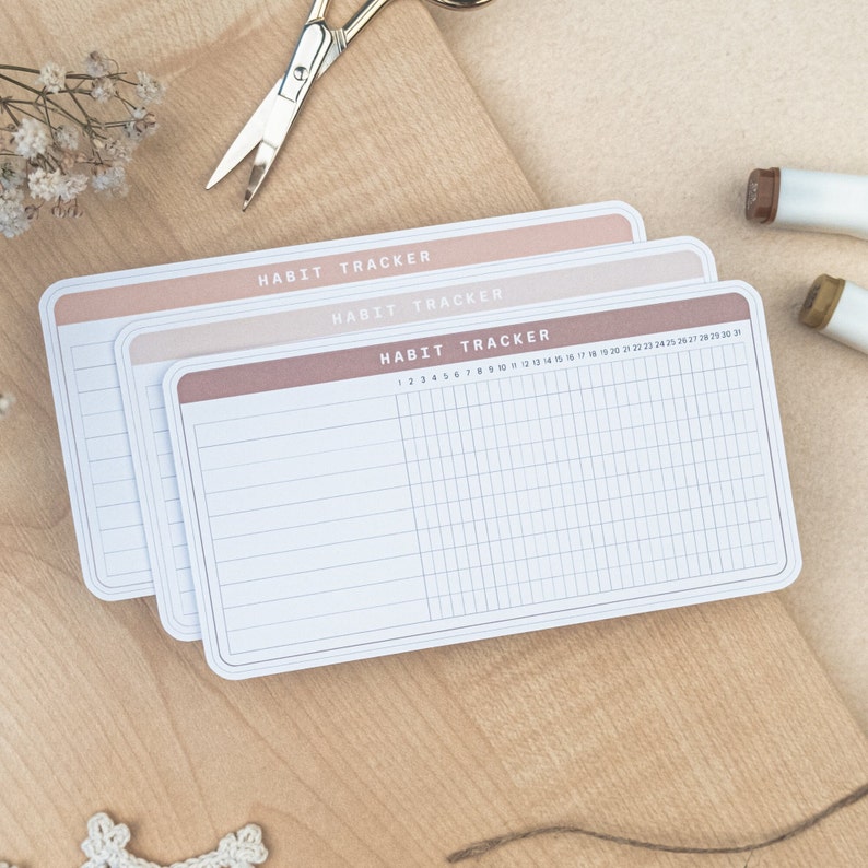 May include: Three habit tracker sheets with a brown, pink, and light pink background. The sheets have a grid pattern and the text "HABIT TRACKER" at the top. The numbers 1 through 31 are printed at the top of the bottom sheet.