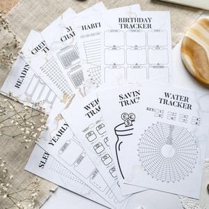 Puede incluir: Un conjunto de hojas de planificación blancas con un patrón de mármol dorado. Las hojas incluyen rastreadores de hábitos, cumpleaños, ahorros y consumo de agua. La palabra "BUJO" es visible en un cuaderno. También hay bolígrafos y elementos decorativos.