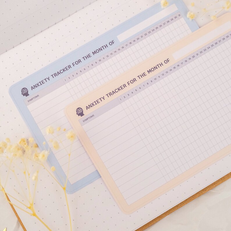 May include: Two printable anxiety tracker sheets with a light blue and cream background. The sheets have a grid pattern and the text "Anxiety Tracker for the Month of" at the top. The text "Symptoms" is below the title.