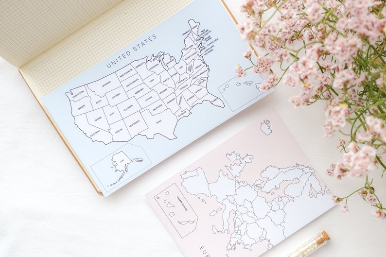 May include: Two map prints, one of the United States and one of Europe, are displayed on a white surface. The US map is light blue with black outlines and the word "UNITED STATES" at the top. The European map is pink with black outlines.