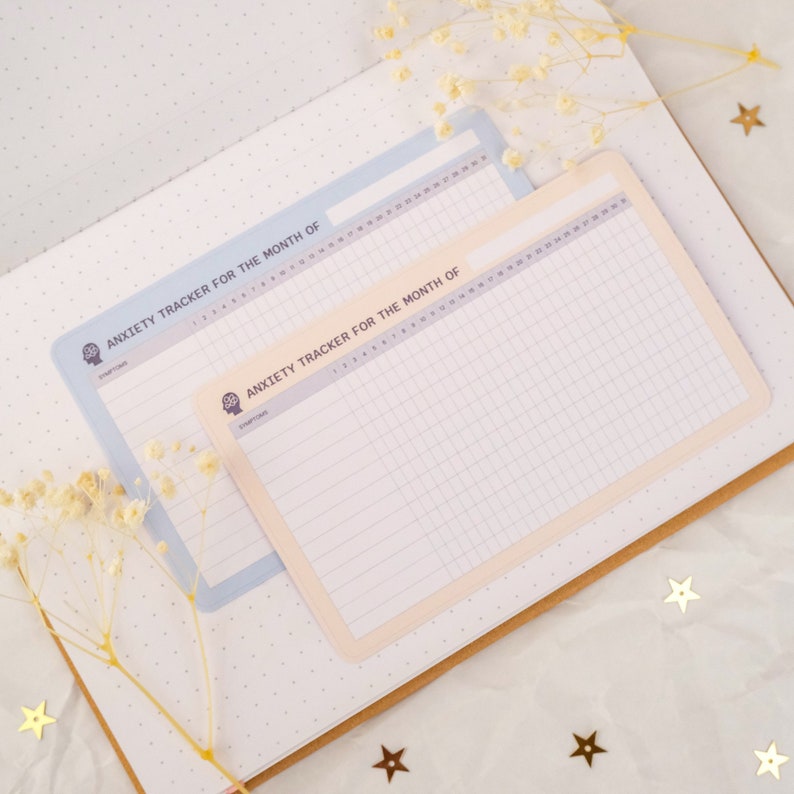 May include: Two printable anxiety trackers for the month of. The trackers are on white paper with a light blue and light peach border. The trackers have a grid pattern and the text "Anxiety Tracker for the Month of" is printed at the top.