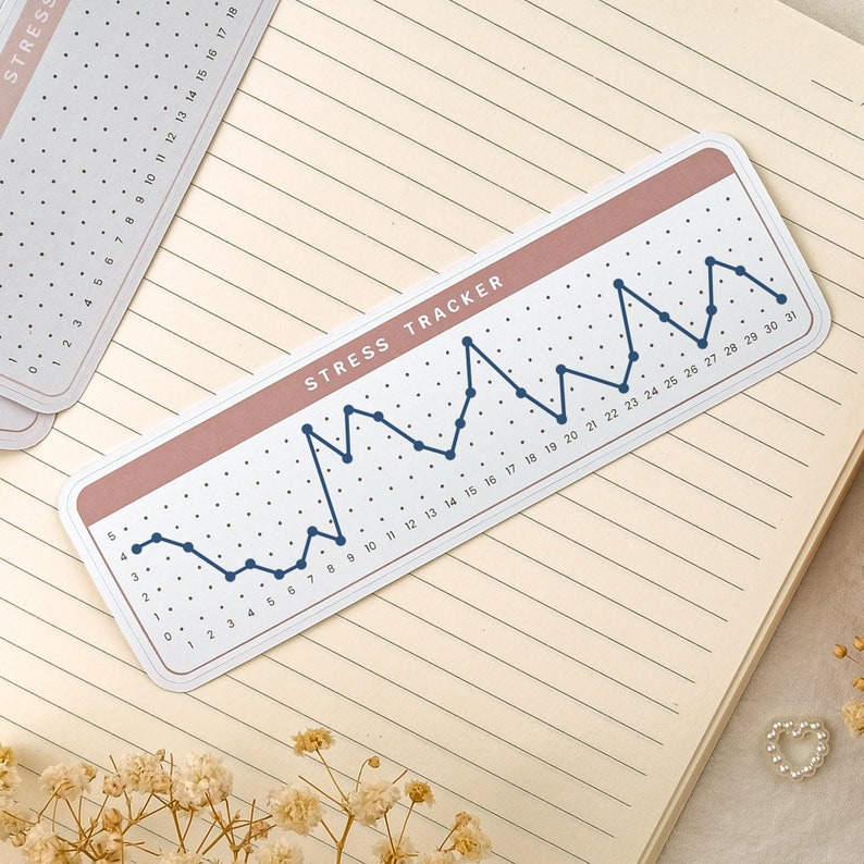 May include: A white sticker with a pink border and a blue line graph. The graph is labeled "Stress Tracker" and has numbers from 1 to 31 along the bottom. The graph shows a person's stress levels over a month.