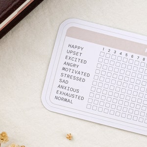 May include: A white card with a mood tracker. The card has a grid with numbers 1 through 8 and a list of emotions: Happy, Upset, Excited, Angry, Motivated, Stressed, Sad, Anxious, Exhausted, and Normal.