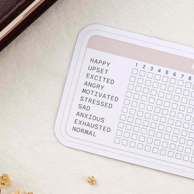 May include: A white card with a mood tracker. The card has a grid with numbers 1 through 8 and a list of emotions: Happy, Upset, Excited, Angry, Motivated, Stressed, Sad, Anxious, Exhausted, and Normal.
