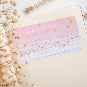 Może przedstawiać: Naklejka do śledzenia snu w kolorze r&oacute;żowym i białym z czarnym wykresem liniowym i tekstem "SLEEP TRACKER" złotymi literami. Naklejka przedstawia p&oacute;łksiężyc i gwiazdy.