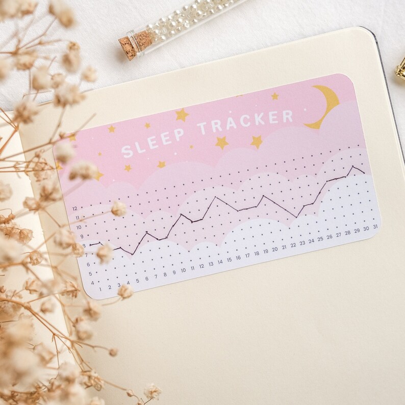 Może przedstawiać: Naklejka do śledzenia snu w kolorze r&oacute;żowym i białym z czarnym wykresem liniowym i tekstem "SLEEP TRACKER" złotymi literami. Naklejka przedstawia p&oacute;łksiężyc i gwiazdy.