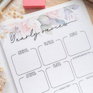 May include: A white journal for yearly review, featuring floral designs and the script text "Yearly review." It includes sections for "Favorite Moments," "Bumps in the Road," and "Accomplishments." A pink eraser and pen are also visible.