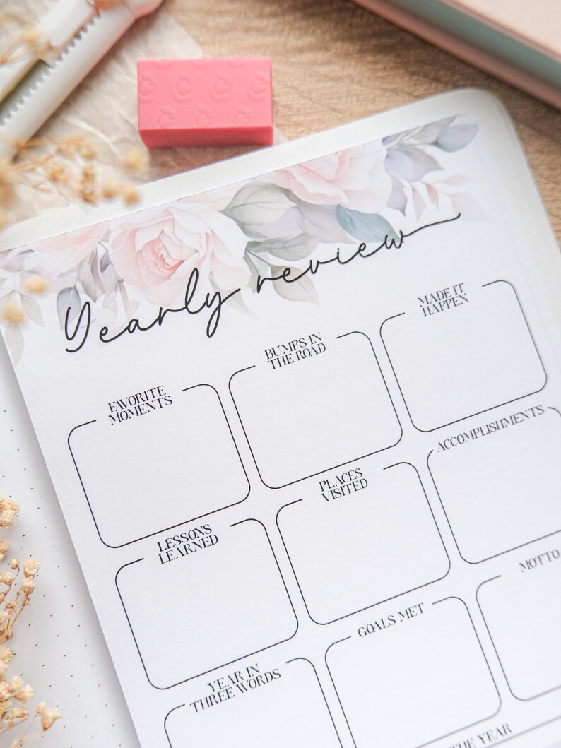 May include: A white journal for yearly review, featuring floral designs and the script text "Yearly review." It includes sections for "Favorite Moments," "Bumps in the Road," and "Accomplishments." A pink eraser and pen are also visible.
