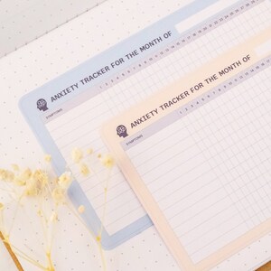 May include: Two printable anxiety trackers with a brain icon and the text "Anxiety Tracker for the Month of" on a white background. The trackers are on a white dotted surface.