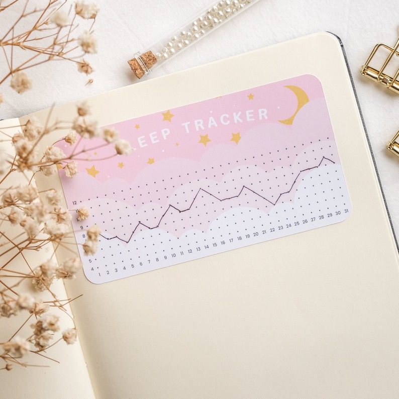 Może przedstawiać: Naklejka do śledzenia snu w kolorze r&oacute;żowym i białym z czarnym wykresem liniowym i tekstem "SLEEP TRACKER" w kolorze białym. Naklejka ma p&oacute;łksiężyc i złote gwiazdki.
