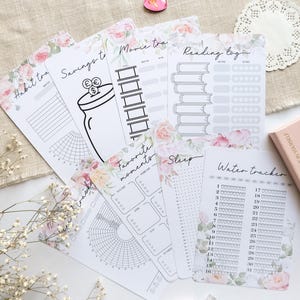 Könnte beinhalten: Eine Sammlung weißer Planer-Einlagen mit floralen Akzenten und verschiedenen Tracking-Layouts, darunter Gewohnheiten, Ersparnisse, Filme, Lesen und Wasser. Ein rosa herzförmiges Objekt und ein Stift sind ebenfalls zu sehen.