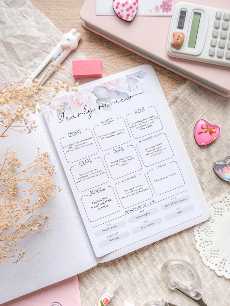 May include: An open planner with a "Yearly Review" page, floral design and sections for notes. A pink notebook, calculator, and decorative items surround the planner. A pen and eraser are also visible.
